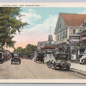 May include: Vintage postcard depicting Main Street in Port Washington, L.I. The image shows a street lined with early 20th-century cars, buildings, and storefronts, including "LA PRIMADORA" and "ELECT". The sky is a mix of blue and light clouds.