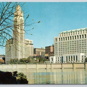 May include: A vintage postcard view of the Columbus, Ohio skyline featuring the LeVeque Tower and the City Hall building situated along the Scioto River. The scene includes a clear blue sky, a calm river, and bare tree branches in the foreground.