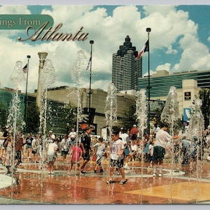 May include: A vintage postcard with the text "Greetings From Atlanta" above a scene of a public fountain with people playing in the water. Buildings and flags are in the background under a blue sky.