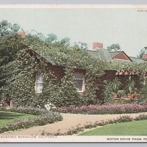 May include: A vintage postcard depicting a rose-covered bungalow in California. The image shows a brick building with a path leading to the entrance, surrounded by lush greenery and flowers. The text reads "A Rose Embowered Bungalow, California."
