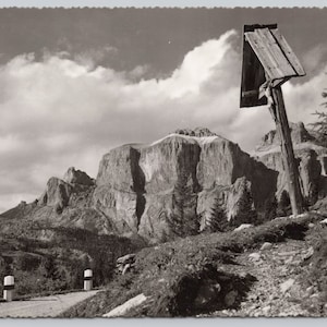 May include: Black and white vintage postcard featuring a mountain landscape with a wooden cross. The cross is on a hillside, with a road and guard posts in the foreground. The sky is cloudy.