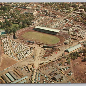 May include: Aerial view of a racetrack with a dirt oval, grandstands, and a large crowd. Surrounding the track are buildings, parking lots, and a river. The image has a vintage postcard aesthetic.