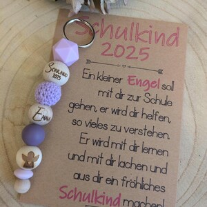 Könnte beinhalten: Ein Schlüsselanhänger mit einem silbernen Ring, einer hellvioletten geometrischen Perle und einer Reihe von Holz- und Häkelperlen. Die Holzperlen sind mit Text und einem Engel graviert. Eine Karte mit deutschem Text lautet "Schulkind 2025".