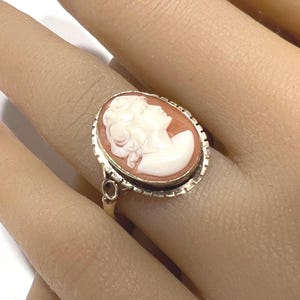 May include: Oval cameo ring featuring a profile of a woman in white against a peach background. The ring has a gold band and a decorative, textured border around the cameo. A small, round accent stone is visible on the band.