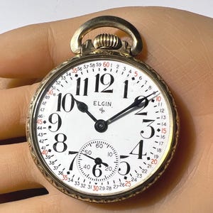 May include: A gold-toned Elgin pocket watch with a white face and black numerals. The watch features a smaller subdial and a second hand. The watch is held in a hand.