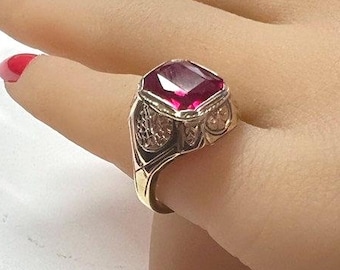 May include: A gold ring featuring a large, square-cut, ruby-red gemstone. The ring has an ornate design with filigree detailing on the sides and a wide band. The gemstone is set in a gold bezel.
