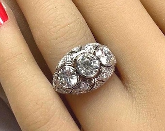 Könnte beinhalten: Nahaufnahme eines silbernen Rings mit drei großen, runden, klaren Steinen in der Mitte, umgeben von kleineren Steinen. Der Ring befindet sich an einem Finger, roter Nagellack ist sichtbar.