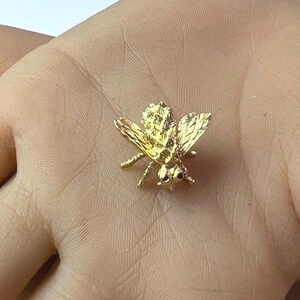 May include: A small, detailed gold-colored bee pin. The bee has textured wings and body, with small black accents for eyes. The pin is likely made of metal and is designed to be worn as jewelry or attached to clothing.
