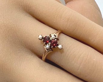 Könnte beinhalten: Ein goldfarbener Ring mit einem Cluster aus roten und weißen Edelsteinen. Das Design des Rings umfasst eine zentrale Anordnung roter Steine, möglicherweise Granate, umgeben von kleineren weißen Steinen, wahrscheinlich Perlen. Der Ring ist auf einem zarten Band gefasst.