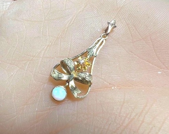 May include: A gold pendant featuring a bow design with a small flower and a round, opalescent stone. The pendant has a delicate, ornate design and is suspended from a small loop.