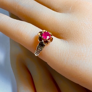 May include: A gold ring with a red ruby gemstone set in a flower design. The ring band is engraved with the word "Gold".