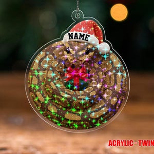 May include: A clear acrylic Christmas ornament featuring a snake design. The snake is brown and tan with a red bow and a Santa hat that says "NAME". The ornament has a twinkle effect and the text "ACRYLIC - TWINKLE".