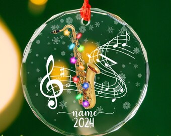 May include: A clear, round Christmas ornament with a saxophone design. The saxophone is adorned with colorful lights and set against a backdrop of musical notes and snowflakes. The text "name 2024" is also visible.