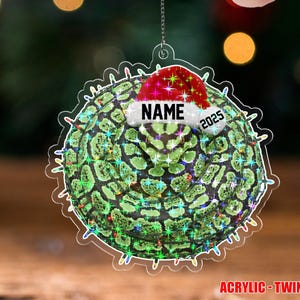 May include: A clear acrylic Christmas ornament with a green and black pattern, a Santa hat, and the text "NAME 2025". The ornament has a string for hanging and is surrounded by colorful lights. The text "ACRYLIC - TWINKLE" is at the bottom.