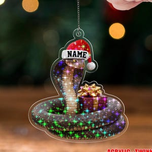 May include: A clear acrylic Christmas ornament featuring a cobra wearing a Santa hat with the word "NAME" on it. The snake is coiled around a gift box with a gold bow. The ornament has a twinkle effect and the text "ACRYLIC - TWINKLE".