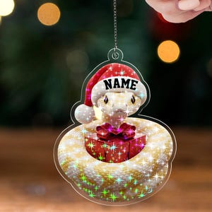 May include: A clear acrylic Christmas ornament featuring a snake wearing a Santa hat with the word "NAME" on it. The snake is coiled around a red gift box with a pink bow. The ornament has a glittery, star-like effect.