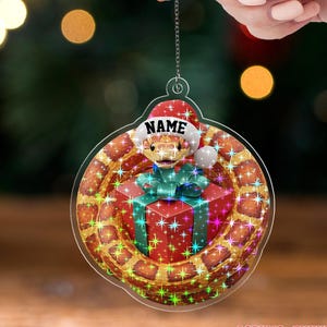 May include: A clear acrylic Christmas ornament featuring a snake wearing a Santa hat with the word "NAME" on it. The snake is coiled around a red gift box with a green bow. The ornament has a chain for hanging and the text "ACRYLIC - TWINKLE".