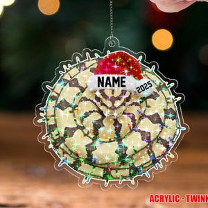 May include: A clear acrylic Christmas ornament with a festive design. The ornament features a snake-like pattern, a Santa hat, and the text "NAME" and "2025". It is trimmed with a string of colorful lights and the words "ACRYLIC - TWINKLE".