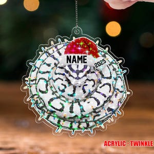 May include: A clear acrylic Christmas ornament with a white and black pattern, a red Santa hat, and the text "NAME 2025". The ornament is surrounded by a string of colorful lights and the text "ACRYLIC - TWINKLE".
