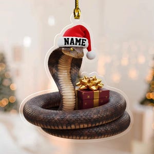 May include: A Christmas ornament featuring a cobra wearing a Santa hat with the word "NAME" on it. The snake is coiled around a wrapped gift box with a gold bow. The ornament hangs from a gold string.