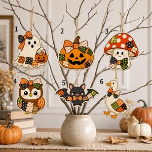 May include: Six wooden hanging ornaments numbered one through six, featuring a ghost, pumpkin, mushroom, owl, bat, and duck. Each character is decorated with colorful patchwork patterns and bows, suspended from a bare branch in a ceramic vase.