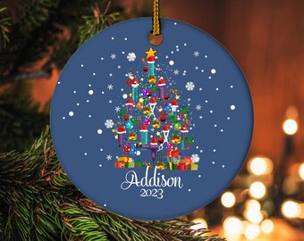 May include: A round, blue Christmas ornament with a gold string. The ornament features a Christmas tree design made of salon tools, ornaments, and gifts. The name "Addison" and the year "2023" are written in white script.