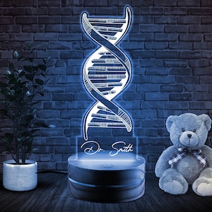 May include: A white wooden night light with a DNA strand design and the text "Dr. Smith" on a clear acrylic panel. The night light is on a wooden base and is illuminated with a white light. A gray teddy bear is sitting on the right side of the image.