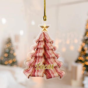 May include: A clear Christmas tree ornament shaped like a tree made of bacon, with a gold star on top and the name "Lucas" in gold glitter script. The ornament hangs from a gold cord.