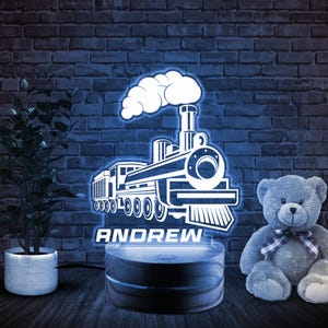 May include: A white wooden night light with a 3D design of a train with the name "ANDREW" underneath. The night light is on a wooden base and is lit up in blue. A gray teddy bear is sitting on the right side of the image.