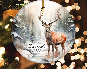 May include: A round, white Christmas ornament with a watercolor painting of a deer standing in a snowy forest. The ornament has the name "David" and the year "2023" written on it.