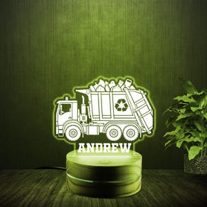 May include: A white silhouette of a garbage truck with a recycling symbol on the side, filled with bags of trash. The truck is on a wooden base with a light-up feature. The name "ANDREW" is written below the truck.