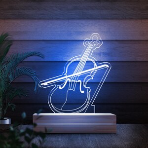May include: A white, illuminated violin-shaped lamp with a bow, set on a rectangular wooden base. The lamp emits a soft, cool blue glow, creating a musical ambiance. The background is a dark wood-paneled wall.