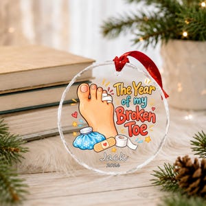 May include: A round, clear glass Christmas ornament with a red ribbon hanger. The design features a cartoon foot with a bandaged toe, a blue ice pack, and a bandage. Text reads: The Year of my Broken Toe, Jack, and 2026.