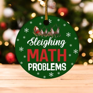 May include: A round, green Christmas ornament with the words "Sleighing MATH PROBLEMS" in white and red. The ornament features a graphic of Santa Claus in a sleigh pulled by reindeer, with snowflakes around the edges.