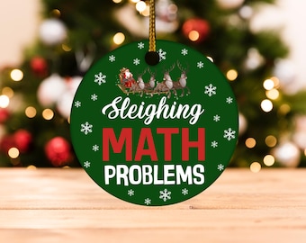 Może przedstawiać: Okrągła, zielona ozdoba świąteczna z napisem "Sleighing MATH PROBLEMS" w kolorze białym i czerwonym. Ozdoba przedstawia Świętego Mikołaja w saniach ciągniętych przez renifery, z płatkami śniegu. Zawieszona na złotym sznurku.