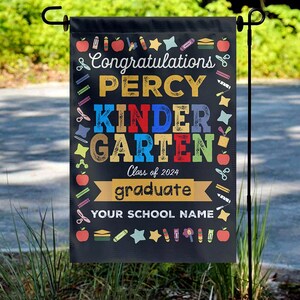 May include: Black garden flag with the text "Congratulations PERCY KINDERGARTEN Class of 2024 graduate YOUR SCHOOL NAME" in colorful letters. The flag is decorated with apples, pencils, stars, and scissors.
