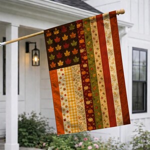 May include: A rectangular garden flag featuring a patchwork design with a dark brown canton filled with orange, red, and green maple leaves. The stripes consist of vertical panels with yellow gingham, autumn foliage, pumpkins, and red stripes.