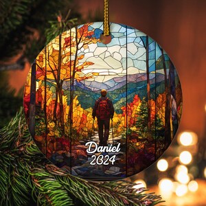 May include: A round Christmas ornament with a stained-glass style design. The image depicts a person hiking on a trail through a colorful forest. The text "Daniel 2024" is written in white script on the bottom of the ornament. A gold string is attached for hanging.