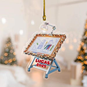 May include: A clear acrylic ornament shaped like a drafting table with a blueprint design. The ornament has a small lamp, string lights, and the text "LUCAS 2025" in red. The ornament hangs from a gold string.