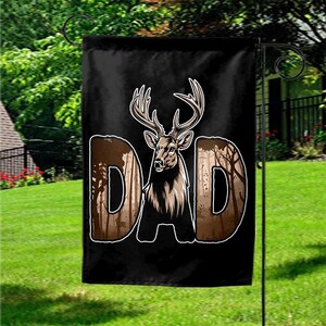 May include: A black garden flag featuring a deer illustration with large antlers. The word "DAD" is displayed in brown letters with a forest scene inside each letter. The flag is hanging on a black metal stand in a grassy yard.