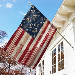 May include: A rectangular decorative flag featuring a patchwork design in a red, white, and blue color scheme. The upper left section displays a dark blue field with a white daisy and red berry floral pattern. The remaining area consists of vertical stripes.
