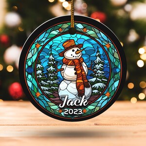 May include: Round Christmas ornament featuring a snowman design. The snowman wears a brown hat and scarf, set against a stained-glass style background with trees and stars. The name "Jack" and the year "2023" are displayed.