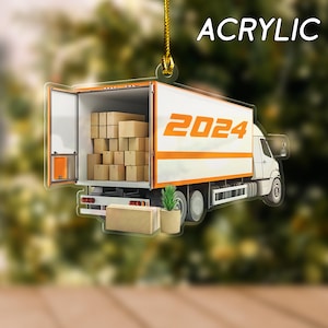 May include: Acrylic ornament featuring a white delivery truck with an orange stripe and the year 2024 in orange. The truck bed is open and filled with cardboard boxes. A small plant in a pot and a cardboard box are in front of the truck.