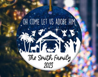 May include: Round Christmas ornament with a blue background and white text and illustrations. The text reads "OH COME LET US ADORE HIM" and "The Smith Family 2023". The ornament features a nativity scene with angels, palm trees, and stars.