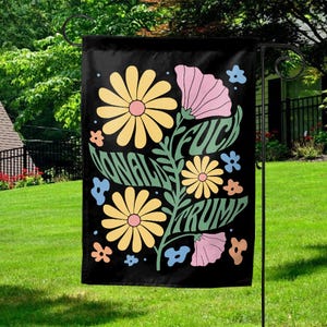 May include: Black garden flag with a floral design and the words "NATIONAL FUCK TRUMP" in a whimsical font. The flowers are yellow, pink, and blue, set against a green stem.