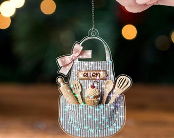May include: A clear acrylic ornament shaped like a baker's apron, with a gray and white striped pattern. It features a pink bow, the name "ellen", and miniature baking tools. The text "ACRYLIC - TWINKLE" is at the bottom.