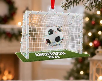 May include: A flat, rectangular Christmas ornament featuring a white soccer goal net with a black and white soccer ball inside. The net is decorated with colorful string lights. The base is green turf with the white text ANDERSON 2025 printed on it.