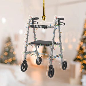 May include: A silver walker Christmas ornament with black wheels and a black seat. The walker is decorated with colorful Christmas lights. The name "SARAH" is printed on the walker.