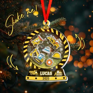 May include: A clear, round Christmas ornament with a construction theme. The ornament features yellow construction vehicles, hard hats, and a gear inside. The bottom of the ornament reads "LUCAS 2025". The ornament is suspended by a red ribbon.