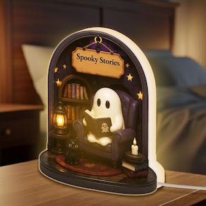 May include: A night light shaped like a spooky scene. A white ghost reads a book in a purple armchair, with a black cat, a lantern, and a sign that says "Spooky Stories". The scene is lit by a candle and small stars.
